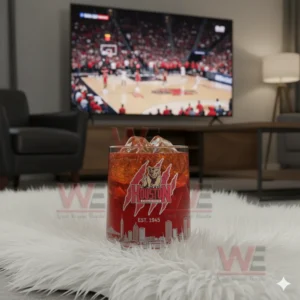 Alt text: A whiskey glass with the Houston Cougars logo and a commemorative anniversary box, with a basketball game playing on the TV in the background.