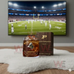 Alt text: A whiskey glass with the Purdue Boilermakers logo and a commemorative anniversary box, with a football game playing on the TV in the background.