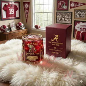 A close view of an anniversary Alabama Crimson Tide glass positioned on a soft rug with sports items surrounding it!