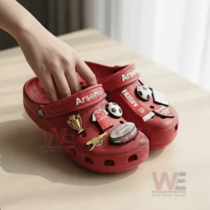 A person adjusting a charm on red Arsenal-themed Crocs with football-related charms.