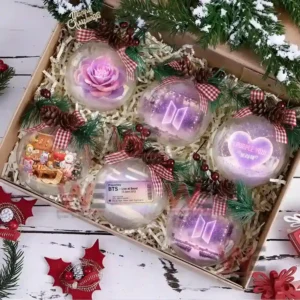 A gift box of BTS themed Christmas ornaments decorated with bows pinecones and snow including designs like a purple flower concert stage heart and ticket.