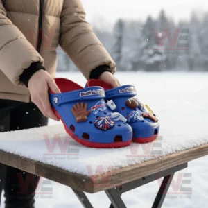 Hands holding Buffalo Bills Crocs decorated with football themed charms over snow covered surface outdoors.