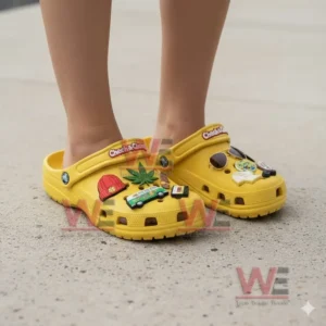 Yellow clogs with multiple decorative charms displayed on feet while standing on a light concrete ground.