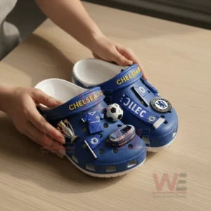 A person adjusting a charm on blue Chelsea FC-themed Crocs decorated with football-related charms.