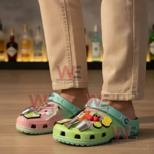 A person wearing pink and green clogs decorated with cocktail charms standing on a wooden floor with bottles behind them!