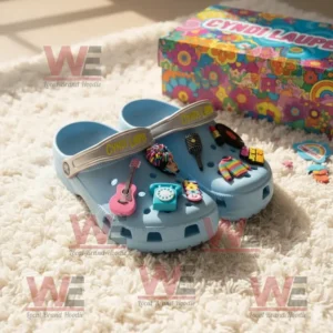 Light blue Crocs with Cyndi Lauper charms including a guitar, microphone, sweater, and record, displayed on a soft carpet beside a colorful patterned box!