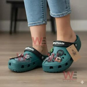 Green clogs with multiple themed charms worn by a person on a wooden floor!