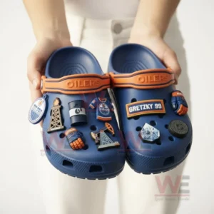Edmonton Oilers Crocs Crocband Clogs 4 A person holding blue Edmonton Oilers Crocs with hockey-themed charms attached.