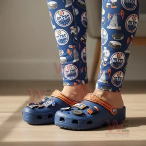 Edmonton Oilers Crocs Crocband Clogs 5 A person wearing blue Edmonton Oilers Crocs decorated with hockey-related charms, paired with Oilers logo leggings.