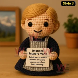 Crochet doll with blond hair in blue dress and gray apron holding Emotional Support Maria sign on wooden table.