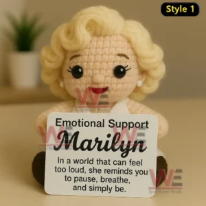 Crochet doll with blonde curls in white dress holding Emotional Support Marilyn sign on light background.
