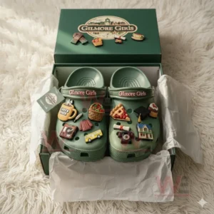 Green Crocs decorated with Gilmore Girls inspired charms like Luke’s Diner mug, slice of pie, graduation cap, and town building shown inside their official green packaging on a white fur surface!
