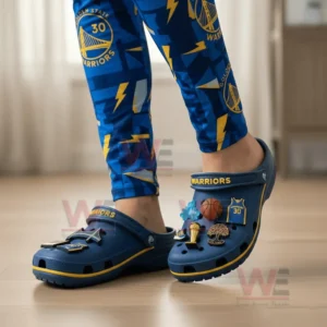 Alt text: A person wearing Golden State Warriors Crocs with themed charms and blue leggings, standing indoors.