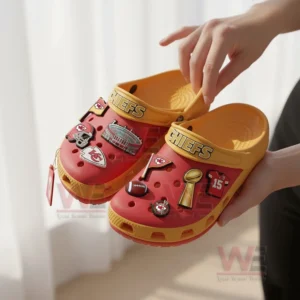 Hands holding a pair of red Crocs with Kansas City Chiefs-themed charms in front of a soft light background.