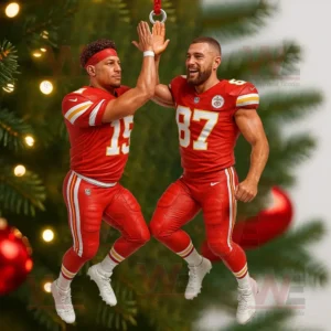 A Christmas ornament of two Kansas City Chiefs football players giving a high five hanging on a green tree with red ornaments and holiday lights in the background!