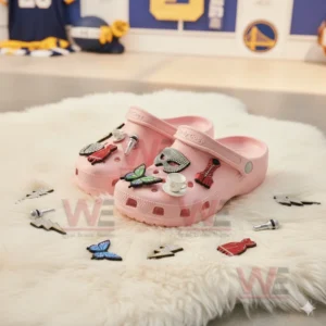 A pair of pink clogs covered in colorful charms displayed on a white furry mat with additional charms placed nearby!