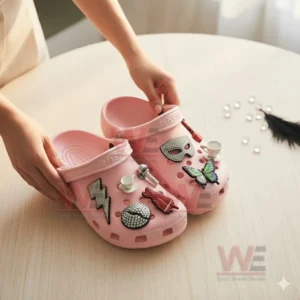 A pair of pink clogs decorated with assorted charms held by hands over a wooden tabletop!