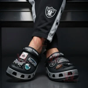 A person wearing Las Vegas Raiders-themed Crocs with skull, football, and dice charms in a dark indoor setting.