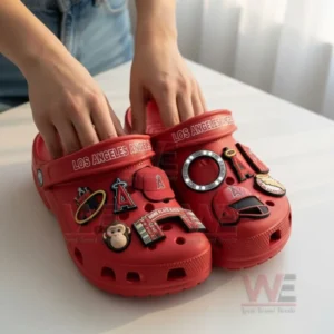 A person holding red Los Angeles Angels Crocs with baseball-related charms and other team-themed accessories.