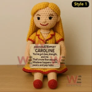 A crochet doll with blonde hair in a yellow dress sitting on a table holding an encouraging message sign!