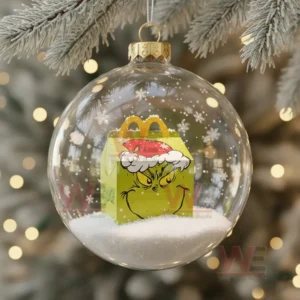 Clear ornament containing a yellow box with a green festive character design sitting on artificial snow!