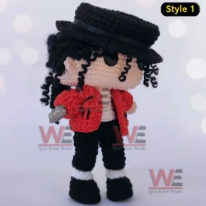 A crochet doll in a red jacket and black hat holding a microphone standing on a plain white surface!