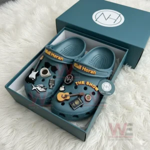 Teal Niall Horan Crocs with musical charms displayed in a branded box on a white fur surface!