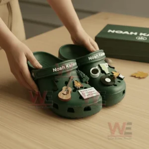 Hands holding green Noah Kahan Crocs with forest and music themed charms on a wood table!