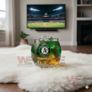 Oakland Athletics whiskey glass with ice and green drink, resting on a rug while a baseball game plays on the TV in the background.