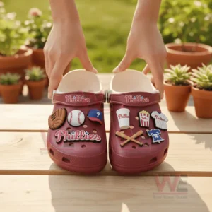 Person holding red Phillies clogs with baseball themed charms outdoors.