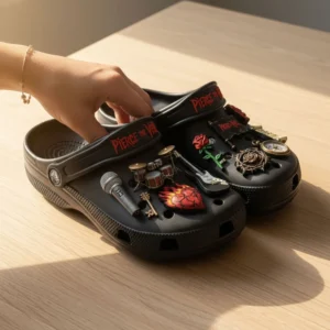 Hand holding Pierce The Veil Crocs with gothic music themed charms on wooden table.