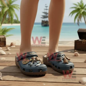 Alt text: A pair of Pirates of the Caribbean Crocs on a wooden deck near the beach with pirate-themed charms on them, and a pirate ship in the background.