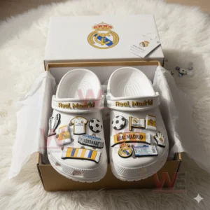 White clogs with Real Madrid charms placed inside an open box resting on a white fluffy rug.