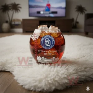 Alt text: A whiskey glass shaped like a baseball with the San Diego Padres logo and ice cubes, placed on a white rug with a baseball game playing on the TV.
