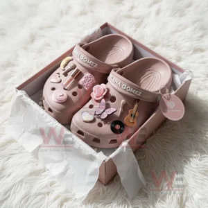Pink clogs with Selena Gomez decorations featuring beauty and music charms resting inside a pink presentation box.