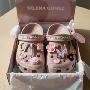 Pink Selena Gomez clogs decorated with musical and beauty charms displayed in a light pink gift box.
