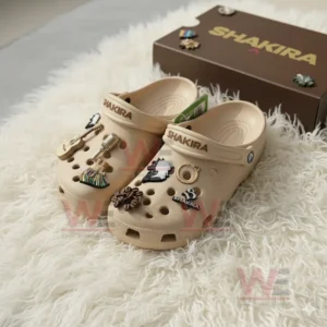 Beige Crocs with Shakira name and musical themed charms displayed beside brown branded box on white furry mat!