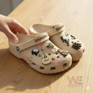 Person holding Shakira Crocs with guitar and microphone charms on wooden table in sunlight!