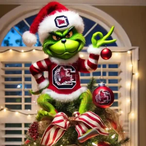 The Grinch dressed in a Santa outfit with a South Carolina Gamecocks logo sitting on top of a Christmas tree holding a red ornament in a brightly lit room!