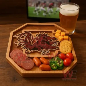 A wooden platter with a Gamecocks design surrounded by assorted snacks sits near a glass of beer with a sports game on a television!