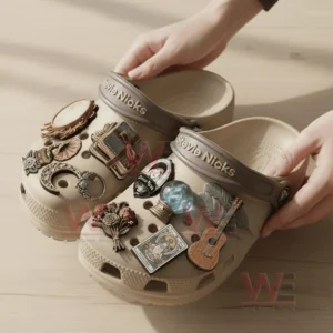 Hands holding beige Stevie Nicks clogs decorated with symbolic and artistic charms on a wooden table.