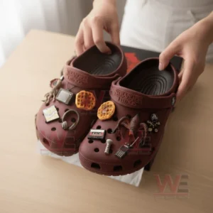 Person holding maroon Stranger Things Crocs with themed charms on wooden table under natural light!