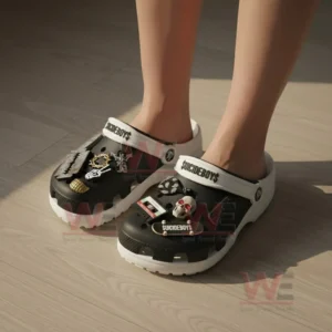 Close-up of a person wearing black and white Suicideboys clogs with metal and skull decorations on a wooden floor.
