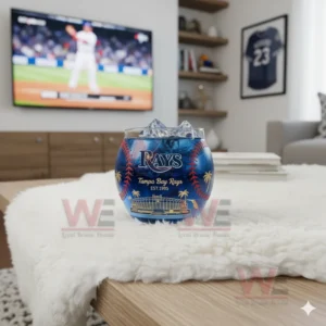 A Tampa Bay Rays whiskey glass with ice on a white rug in front of a television showing a baseball game.