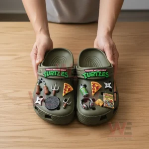Hands holding olive green Teenage Mutant Ninja Turtles Crocs with various themed charms on a wooden table.