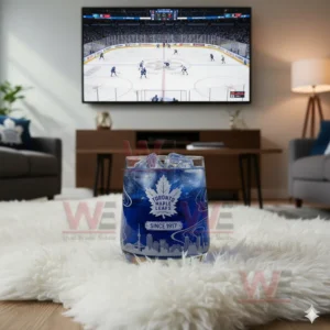 A Toronto Maple Leafs whiskey glass with ice on a white rug in front of a television showing a hockey game.