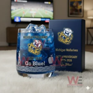 Alt text: A whiskey glass with the Michigan Wolverines logo and a commemorative anniversary box, with a football game playing on the TV in the background.