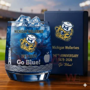 Alt text: A close-up of a whiskey glass with the Michigan Wolverines logo and a commemorative anniversary box, set against a football field backdrop.