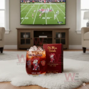 Alt text: A whiskey glass with the Ole Miss Rebels logo and a commemorative anniversary box, with a football game playing on the TV in the background.