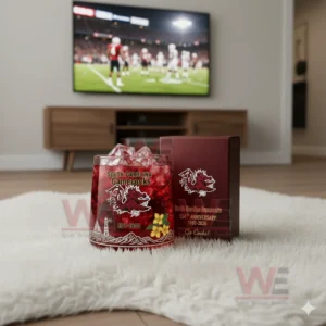 Alt text: A whiskey glass with the South Carolina Gamecocks logo and a commemorative anniversary box, with a football game playing on the TV in the background.